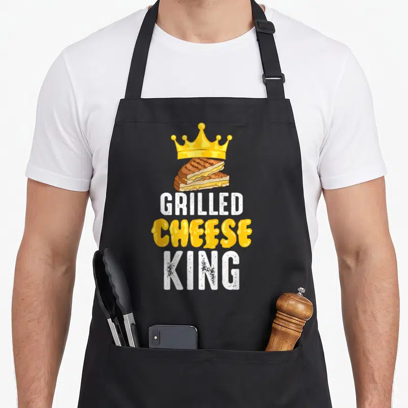 Grilled maker apron - Crown atop toasted sandwich with stacked GRILLED CHEESE KING text in yellow and white — shown on a black cotton canvas apron against a white background