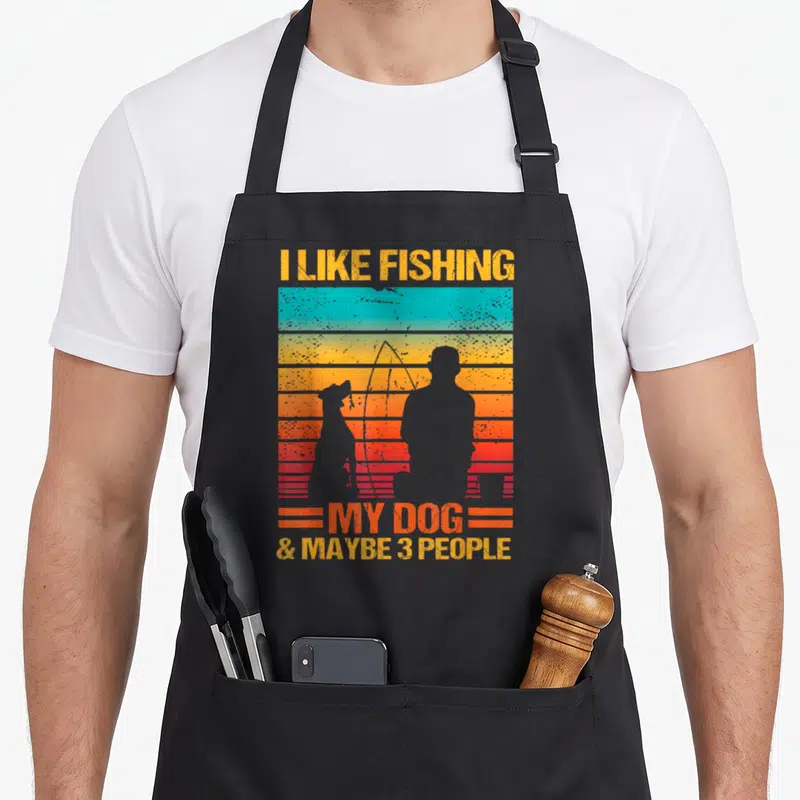 Fishing dog apron - Retro sunset graphic with fisherman and dog silhouettes and text I LIKE FISHING MY DOG & MAYBE 3 — shown on a black cotton canvas apron against a white background