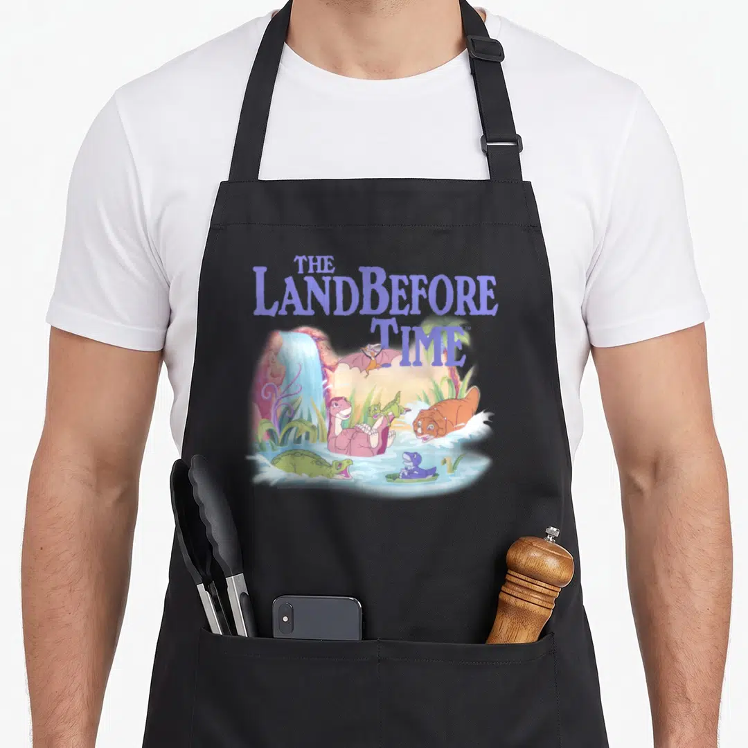 Land dinosaur apron - The Land Before Time title with five cartoon dinosaurs swimming near a waterfall — shown on a black cotton canvas apron against a white background