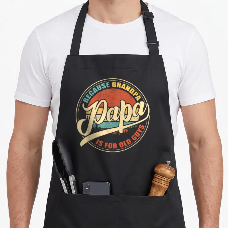 Papa sunset grandpa apron - Retro striped sun with forest silhouette and Because Grandpa Papa Is For Old Guys text — shown on a black cotton canvas apron against a white background