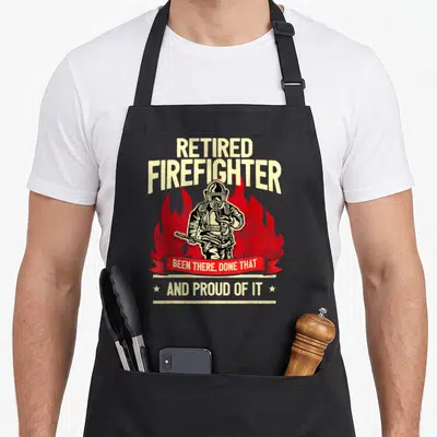 Fire retirement apron - Distressed RETIRED FIREFIGHTER text with red flames and helmeted figure holding hose — shown on a black cotton canvas apron against a white background