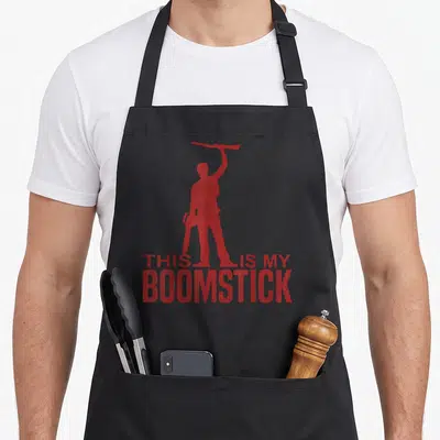 Boomstick chainsaw apron - Red silhouette of man holding shotgun and chainsaw above THIS IS MY BOOMSTICK text — shown on a black cotton canvas apron against a white background