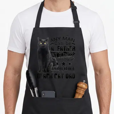Someone cat apron - Black cat sitting beside stacked distressed text reading Any Man Can Be A Father But It Takes — shown on a black cotton canvas apron against a white background