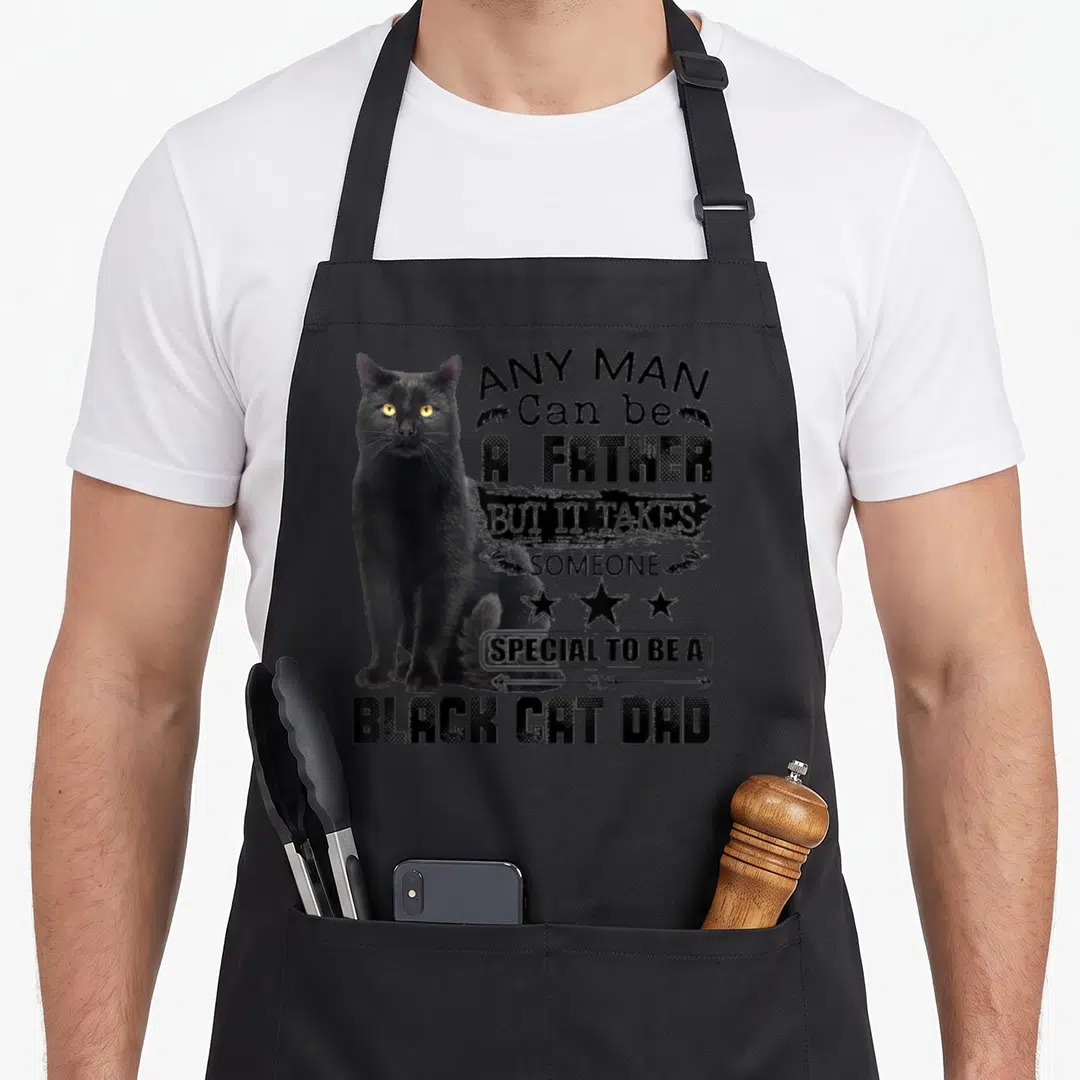Someone cat apron - Black cat sitting beside stacked distressed text reading Any Man Can Be A Father But It Takes — shown on a black cotton canvas apron against a white background