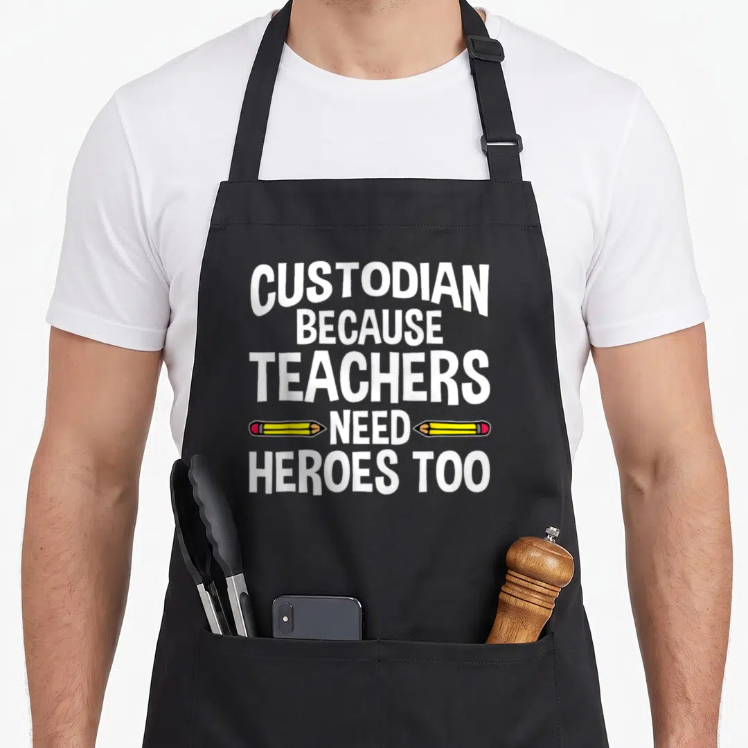 Cute school janitor apron - White text Custodian Because Teachers Need Heroes Too with two yellow pencils flanking NEED — shown on a black cotton canvas apron against a white background