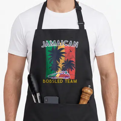 Jamaican summer aprons - Retro bobsled on green yellow red stripes with two palm trees and Jamaican text — shown on a black cotton canvas apron against a white background