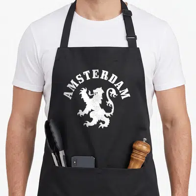 Netherland crest lion apron - White distressed heraldic lion with arched AMSTERDAM text on black fabric — shown on a black cotton canvas apron against a white background