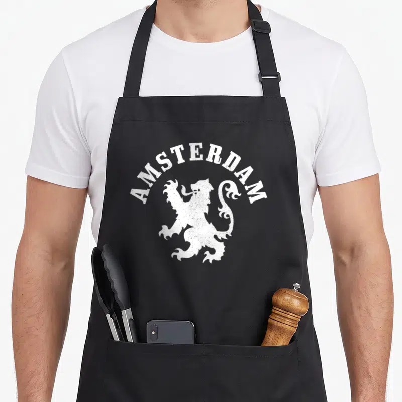 Netherland crest lion apron - White distressed heraldic lion with arched AMSTERDAM text on black fabric — shown on a black cotton canvas apron against a white background