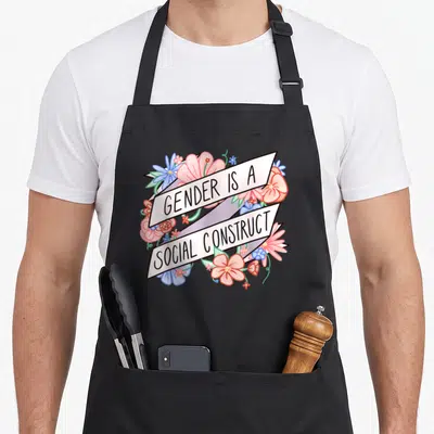 Gender floral apron - Pink and blue flowers surrounding diagonal banners reading Gender Is A Social Construct in black — shown on a black cotton canvas apron against a white background