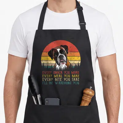 Snack boxer dog apron - Retro boxer dog head before striped sun with trees and four lines of text about watching snacks — shown on a black cotton canvas apron against a white background
