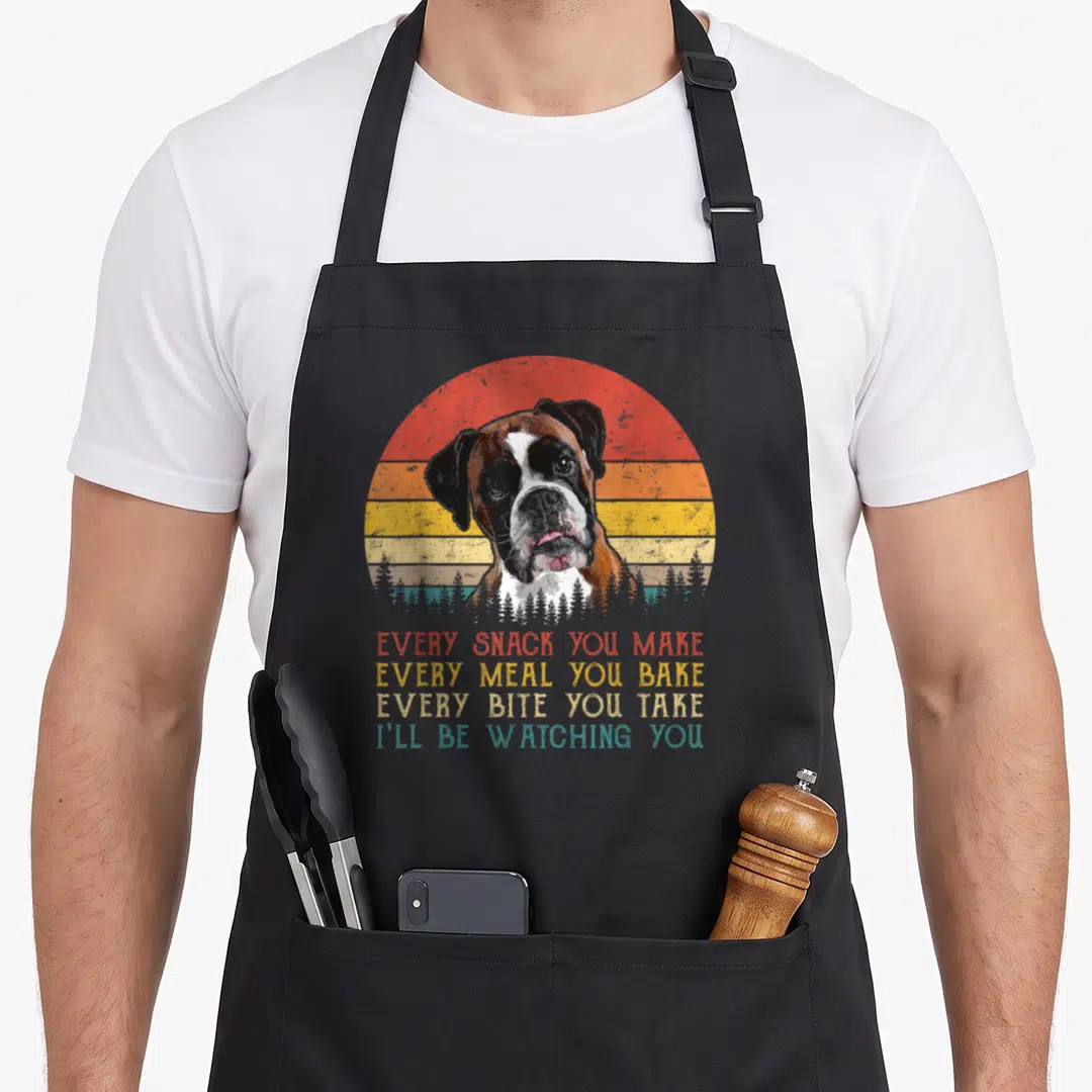 Snack boxer dog apron - Retro boxer dog head before striped sun with trees and four lines of text about watching snacks — shown on a black cotton canvas apron against a white background