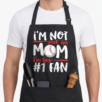His mother's day aprons - Stacked text I'M NOT just his MOM I'm his #1 FAN with red bats and central baseball — shown on a black cotton canvas apron against a white background