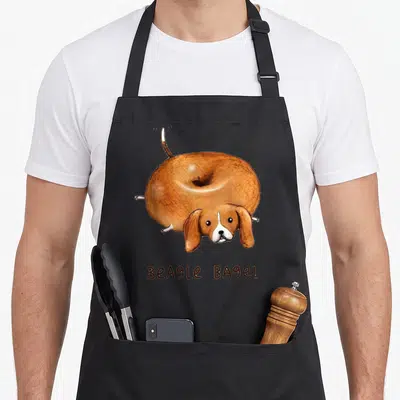 Bagel beagle apron - Cartoon brown and white beagle shaped like a bagel with text below — shown on a black cotton canvas apron against a white background