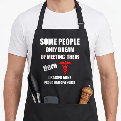 Raised nurses apron - Stacked white text reading I Raised Mine with a red caduceus symbol beside the word Hero — shown on a black cotton canvas apron against a white background