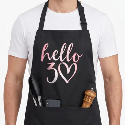 Hello birthday apron - Pink hello text above 30 with heart zero on black bib apron — shown on a black cotton canvas apron against a white background