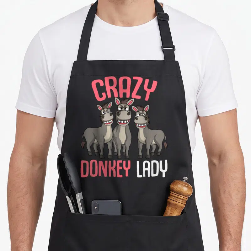 Donkey owner funny apron - Three cartoon donkeys with pink CRAZY DONKEY LADY text — shown on a black cotton canvas apron against a white background