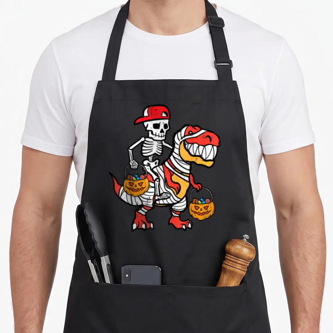 Skeleton mummy and me aprons - Cartoon skeleton in red cap rides bandaged dinosaur holding two orange pumpkin candy — shown on a black cotton canvas apron against a white background