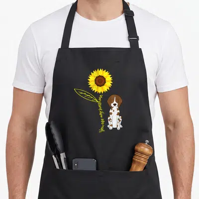 Lovely german apron - Cartoon dog sitting next to yellow sunflower with vertical text reading You are my sunshine — shown on a black cotton canvas apron against a white background