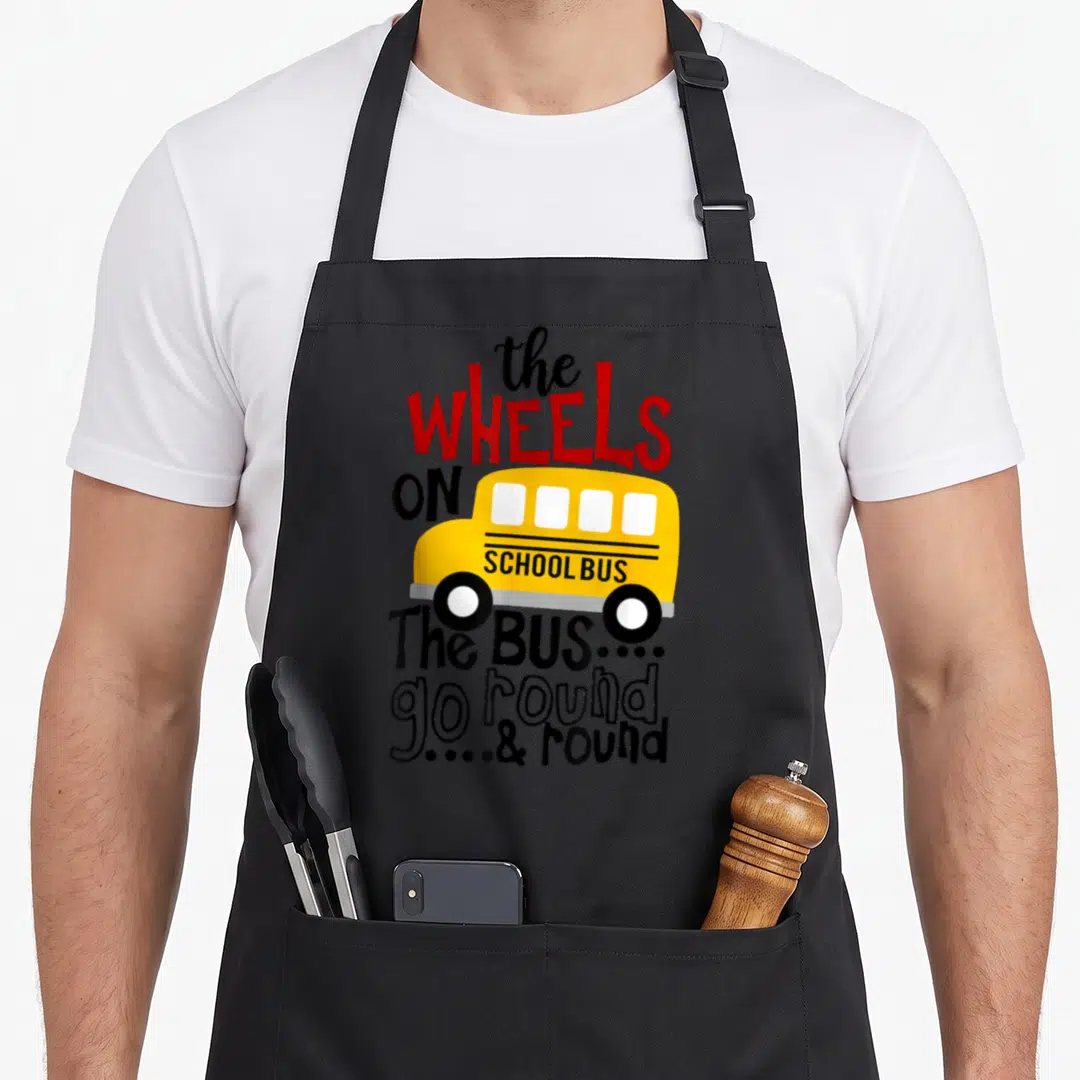 Bus wheel apron - Cartoon yellow school bus with red and black text reading The Wheels On School Bus go round and round — shown on a black cotton canvas apron against a white background