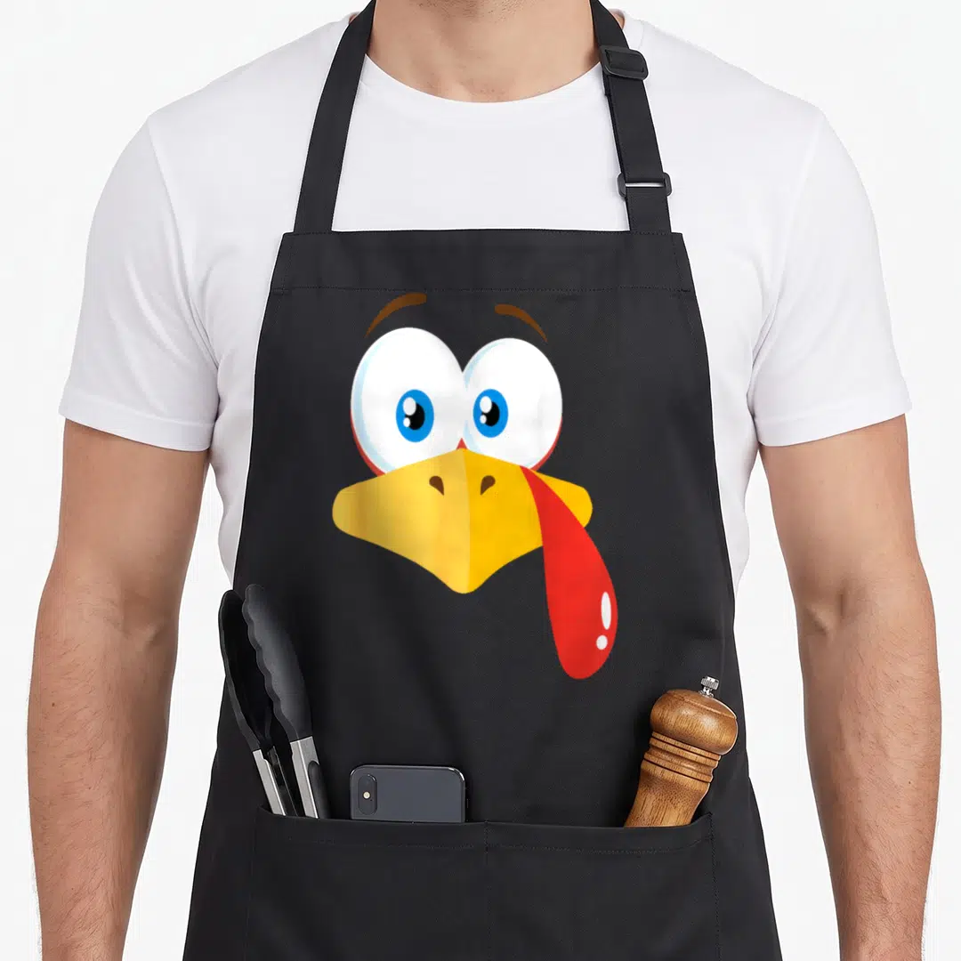 Turkey pilgrim apron - Cartoon bird head with yellow beak, red tongue from side, blue eyes, brown eyebrows — shown on a black cotton canvas apron against a white background
