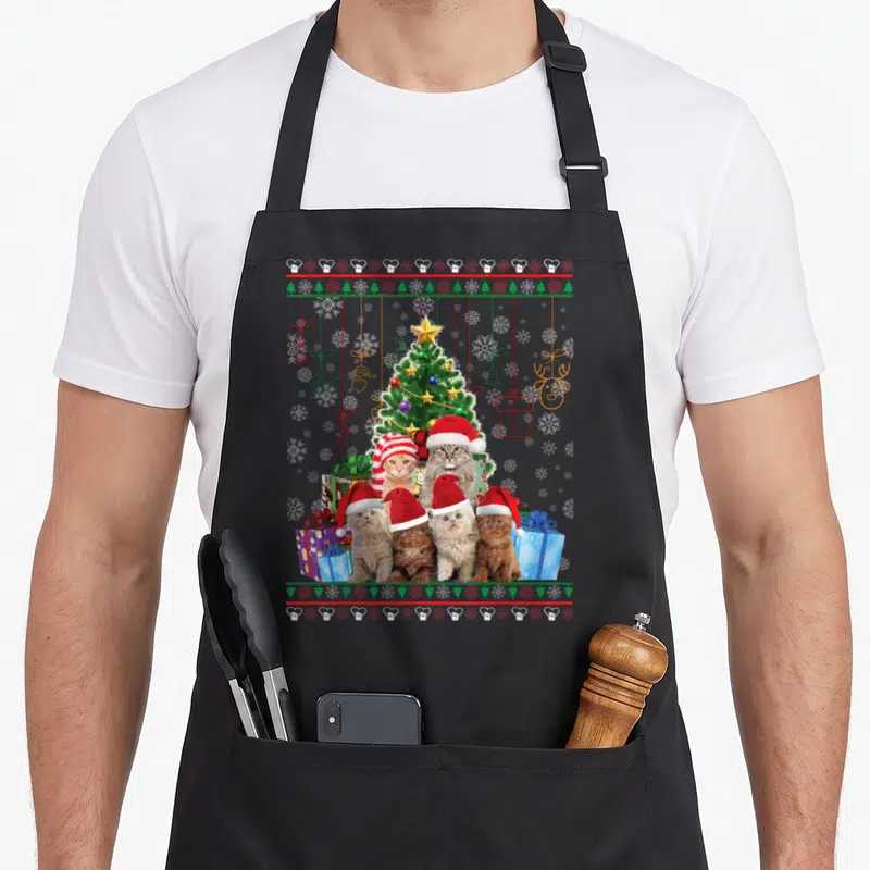 Merry ugly christmas apron - Five cats in Santa hats in front of a Christmas tree with red green and white snowflake — shown on a black cotton canvas apron against a white background