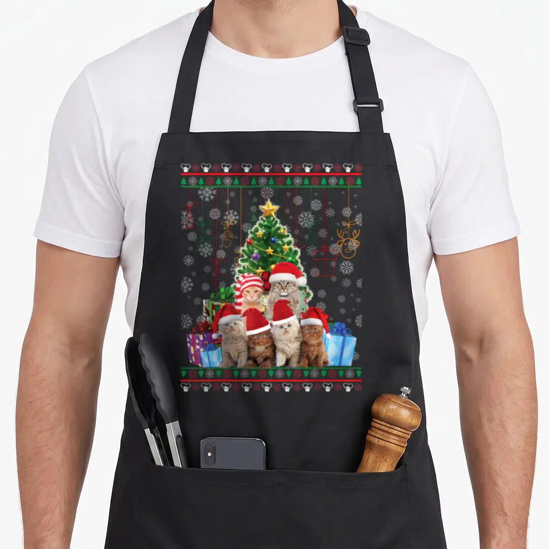 Merry ugly christmas apron - Five cats in Santa hats in front of a Christmas tree with red green and white snowflake — shown on a black cotton canvas apron against a white background