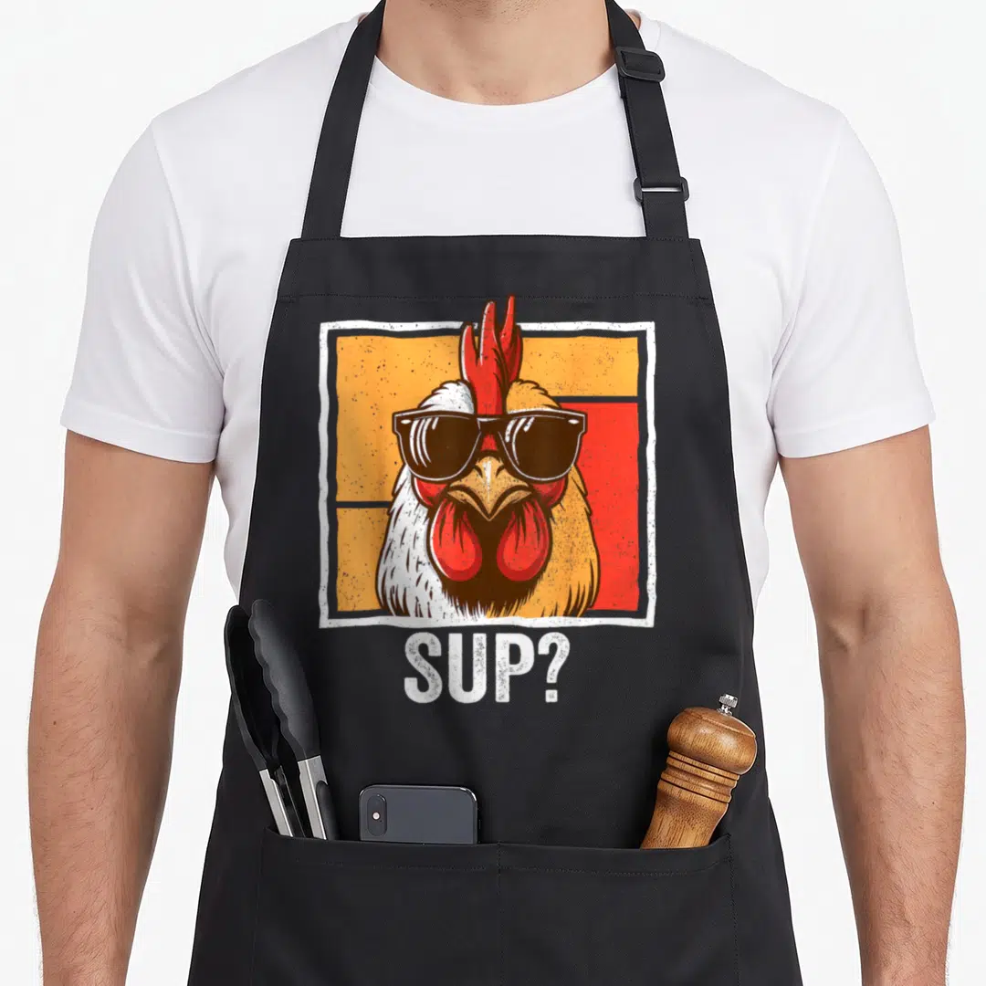 Sunglass farmers apron - Cartoon chicken wearing black sunglasses below white SUP? text on orange and red background — shown on a black cotton canvas apron against a white background