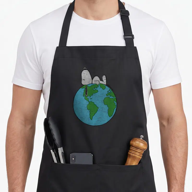Laying earth apron - Snoopy lying on a blue and green globe illustration — shown on a black cotton canvas apron against a white background