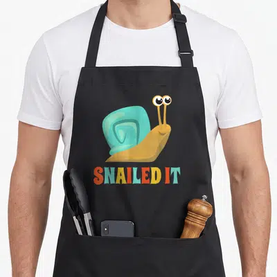 Funny snailed snail apron - Cartoon snail with teal shell above SNAILED IT text in orange, yellow, and white — shown on a black cotton canvas apron against a white background