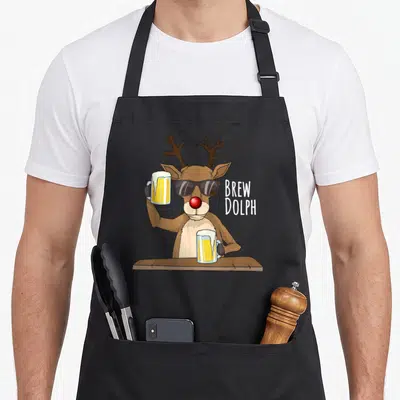Alcohol christmas apron - Cartoon brown reindeer in sunglasses holding two yellow beer mugs with white text BREW DOLPH — shown on a black cotton canvas apron against a white background