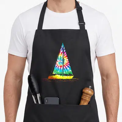 Sailing tie-dye apron - Cartoon sailboat with tie-dye sail and orange water ripples on black fabric — shown on a black cotton canvas apron against a white background
