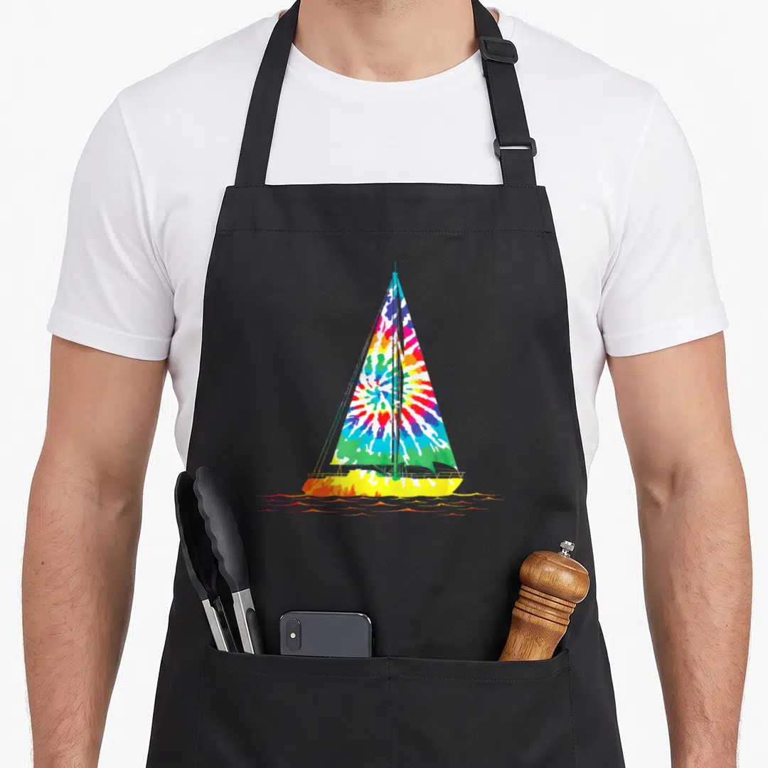 Sailing tie-dye apron - Cartoon sailboat with tie-dye sail and orange water ripples on black fabric — shown on a black cotton canvas apron against a white background