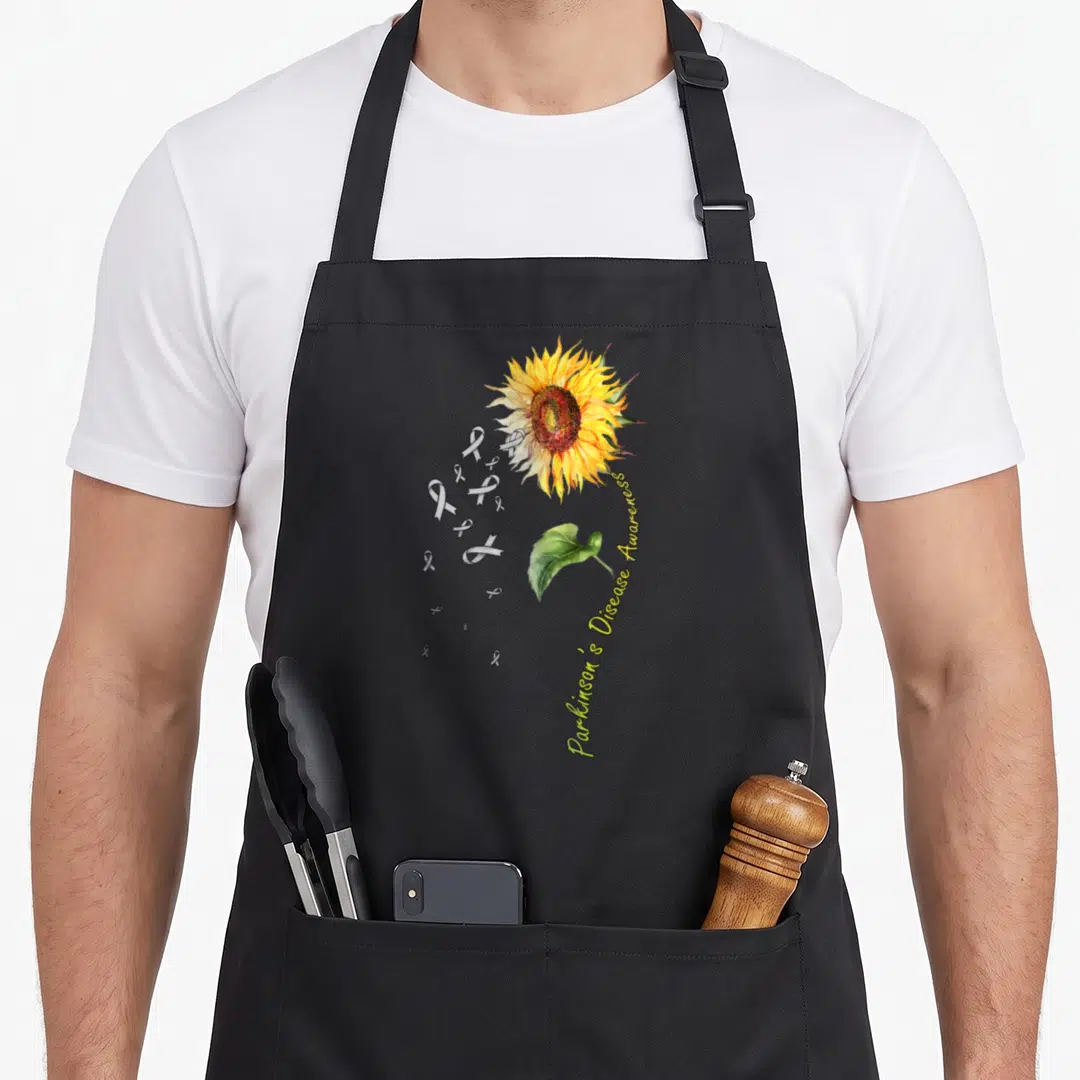 Parkinson sunflower apron - Watercolor sunflower with grey ribbons and green leaf on grey background — shown on a black cotton canvas apron against a white background