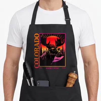 Moose 80s apron - Retro moose in sunglasses with vertical COLORADO text and sunset forest background in red orange — shown on a black cotton canvas apron against a white background