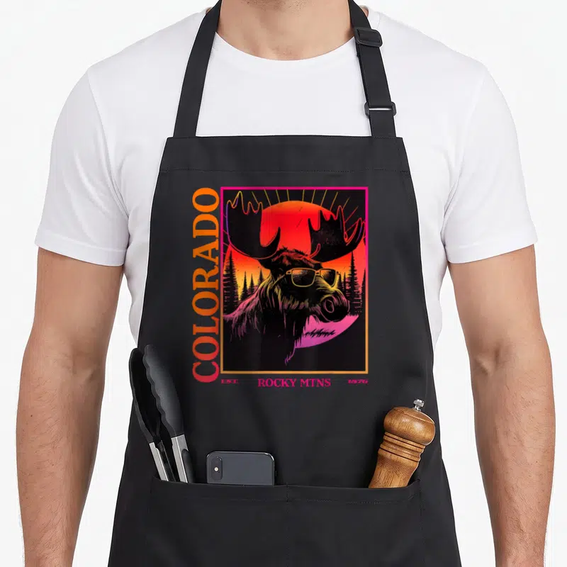 Moose 80s apron - Retro moose in sunglasses with vertical COLORADO text and sunset forest background in red orange — shown on a black cotton canvas apron against a white background