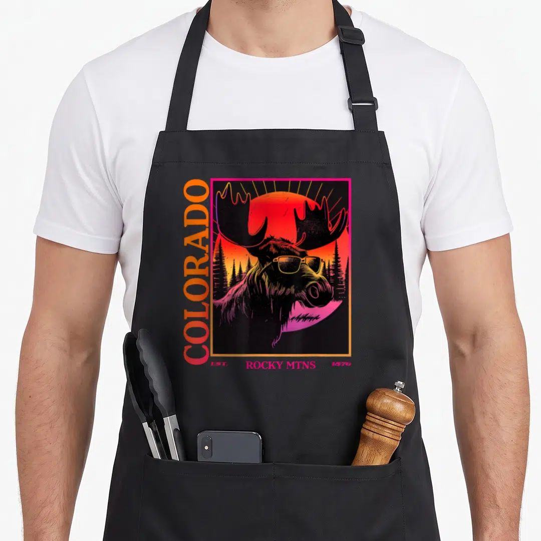 Moose 80s apron - Retro moose in sunglasses with vertical COLORADO text and sunset forest background in red orange — shown on a black cotton canvas apron against a white background
