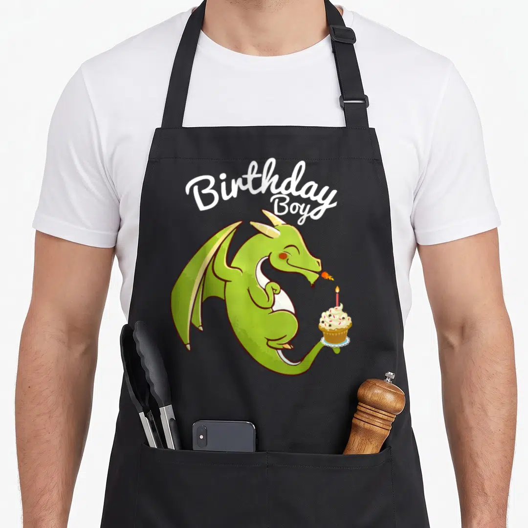 Dragon cupcake apron - Green dragon holding cupcake with lit candle under white Birthday Boys script text — shown on a black cotton canvas apron against a white background