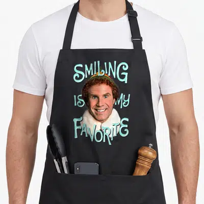 Movie elf apron - Buddy face with arched Smiling Is My Favorite text and black swirls — shown on a black cotton canvas apron against a white background