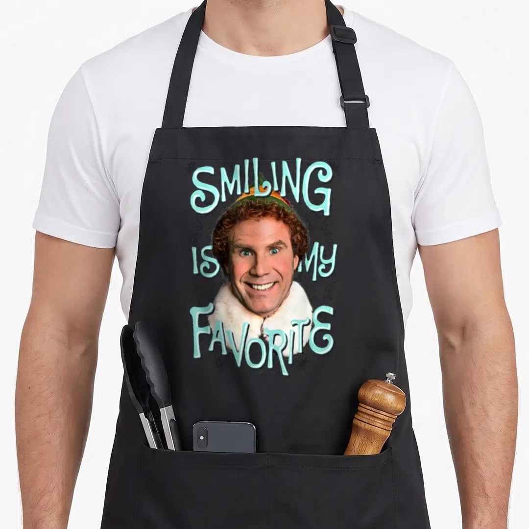 Movie elf apron - Buddy face with arched Smiling Is My Favorite text and black swirls — shown on a black cotton canvas apron against a white background