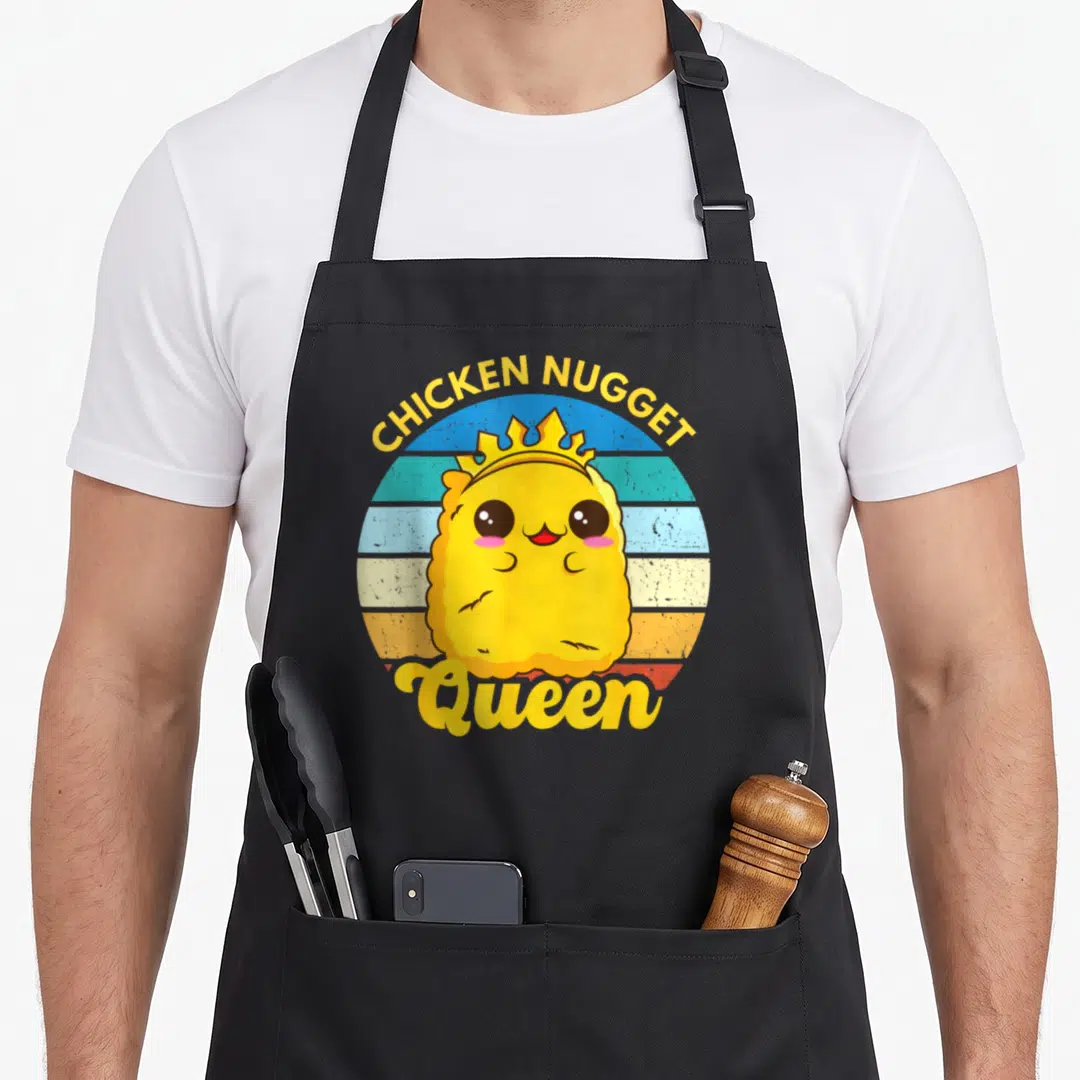 Queen chicken print apron - Yellow cartoon chicken nugget wearing a crown over striped circle with text — shown on a black cotton canvas apron against a white background
