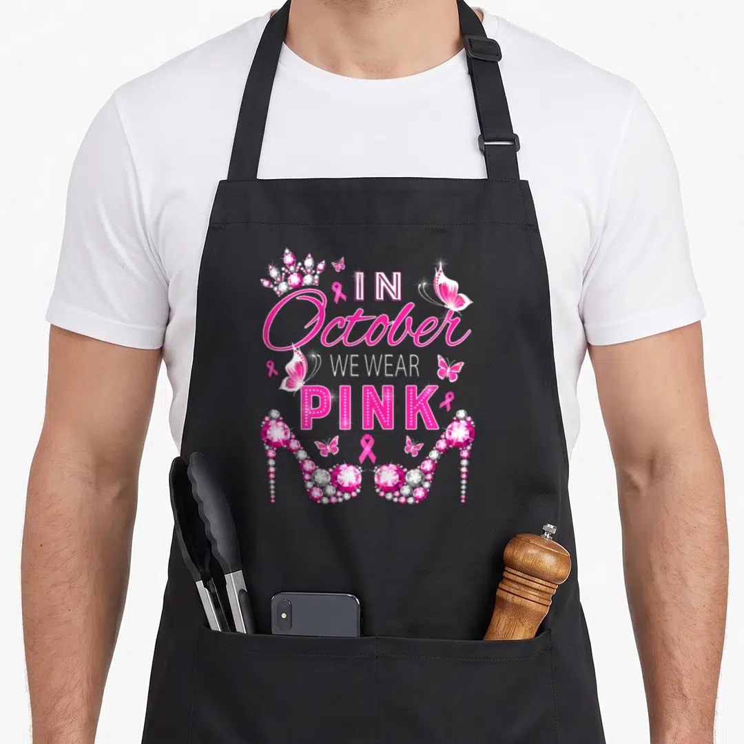 Band breast cancer awareness aprons - IN October WE WEAR PINK text with jeweled heels crown and butterflies — shown on a black cotton canvas apron against a white background