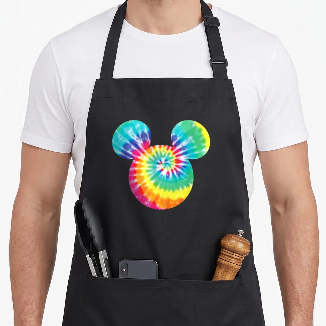 Icon tie dye apron - Mouse head silhouette filled with tie-dye spiral pattern in red, yellow, green, blue, and purple — shown on a black cotton canvas apron against a white background