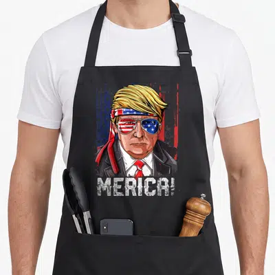 Trump 4th of july apron - Cartoon Trump portrait with flag headband sunglasses and MERICA! text on black apron — shown on a black cotton canvas apron against a white background