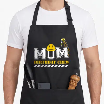Birthday crew aprons - MUM text with yellow hard hat on U, crane hook on M, and BIRTHDAY CREW below — shown on a black cotton canvas apron against a white background