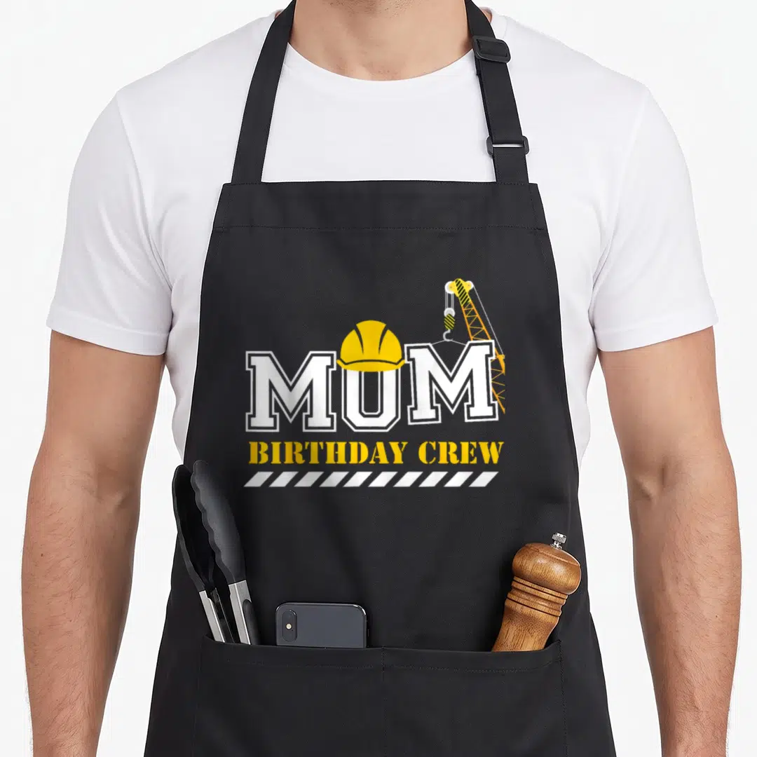 Birthday crew aprons - MUM text with yellow hard hat on U, crane hook on M, and BIRTHDAY CREW below — shown on a black cotton canvas apron against a white background