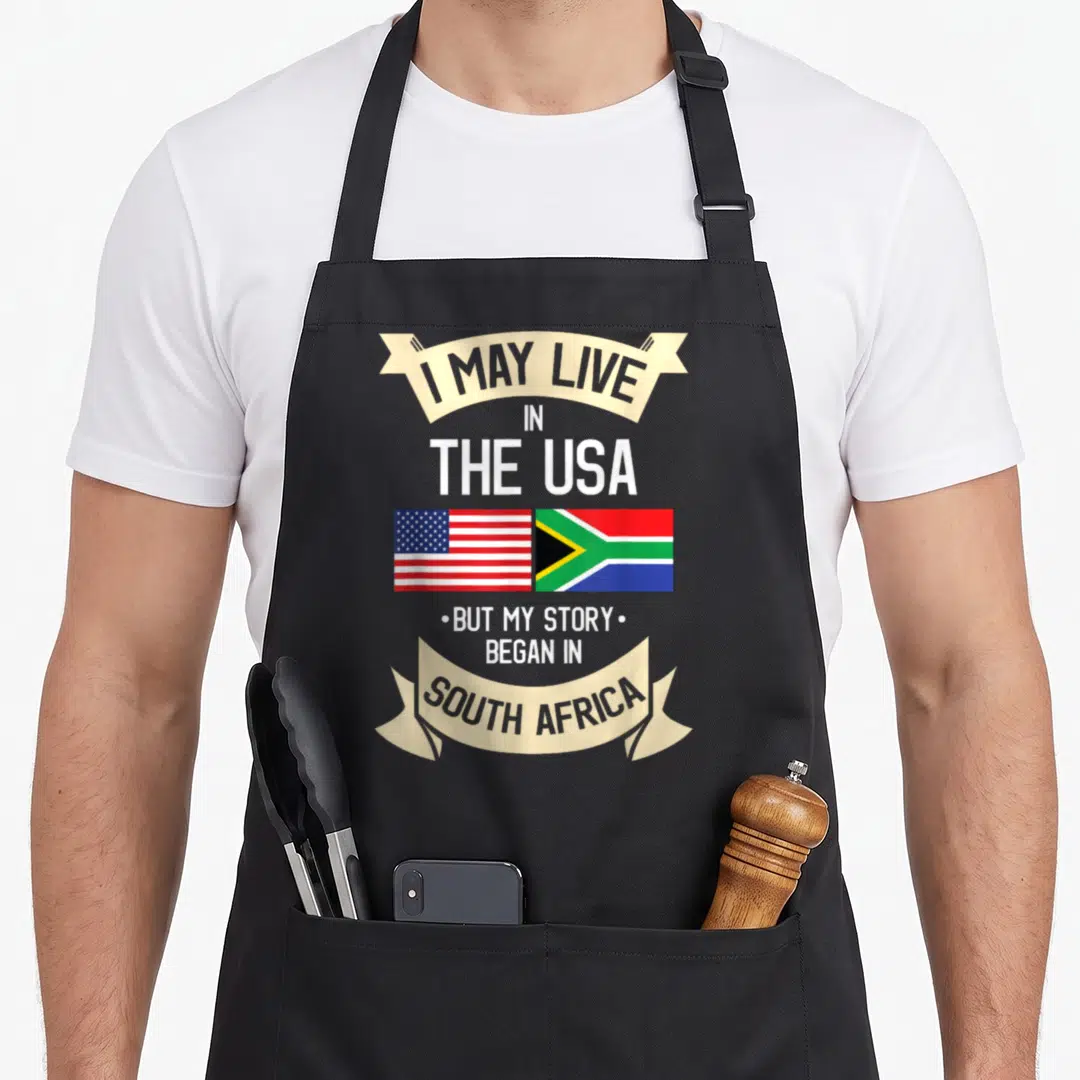 Africa american flag apron - Stacked text I MAY LIVE IN THE USA SOUTH AFRICA with USA and South Africa flags side — shown on a black cotton canvas apron against a white background