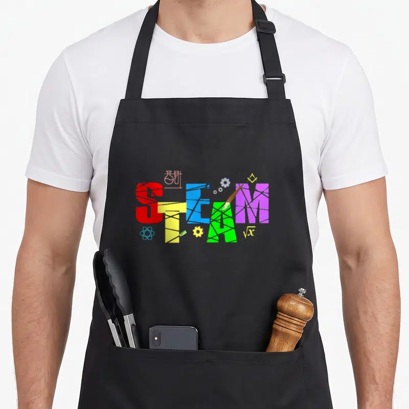 Creativity science apron - Colorful cracked STEAM text with red yellow blue green purple letters and academic icons — shown on a black cotton canvas apron against a white background