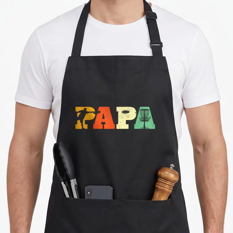 Father golf apron - PAPA text in orange red cream green with disc golf player disc and basket silhouettes — shown on a black cotton canvas apron against a white background