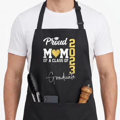 Proud graduate mom class apron - Stacked white text proud mom of class of 2023 graduate with gold heart and cap icon — shown on a black cotton canvas apron against a white background
