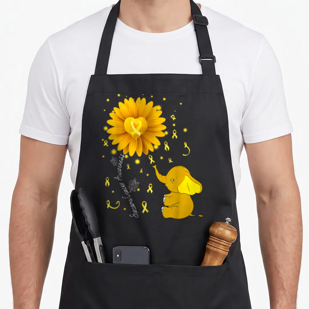 Ribbon daisy apron - Yellow daisy with heart center, cartoon elephant, and Sarcoma Cancer Awareness text on black — shown on a black cotton canvas apron against a white background